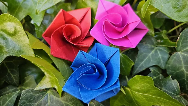 Origami-Rosen in Rot, Rosa und Blau zwischen üppigem grünem Laub platziert.